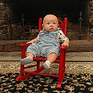 Joseph is registered to the contest to win money with this photo: baby, child, rocking_chair, denim_overalls, socks, indoor, fireplace, stone_wall, rug, expression, seated, furniture, home, curious, infant, clothing, patterned_shirt, wood, floor, portrait
