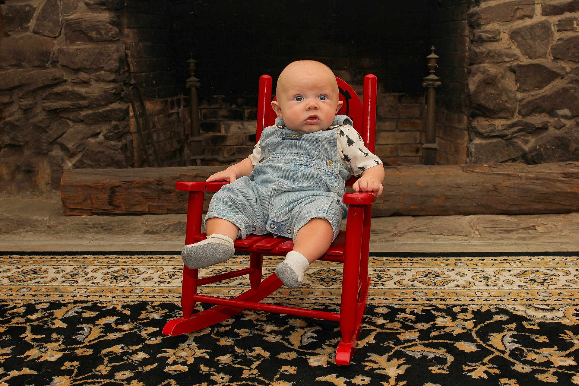 Joseph is registered to the contest to win money with this photo: baby, child, rocking_chair, denim_overalls, socks, indoor, fireplace, stone_wall, rug, expression, seated, furniture, home, curious, infant, clothing, patterned_shirt, wood, floor, portrait