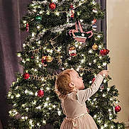 Finnley joined the competition — help win amazing prizes! toddler, child, christmas_tree, ornaments, lights, holiday, indoor, carpet, curtains, decorations, festive, dress, socks, person, celebration, home, seasonal, young_child, curious, standing