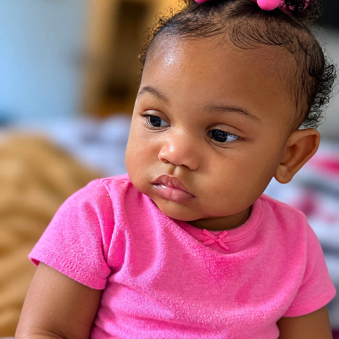 Amariana is registered to the contest to win money with this photo: baby, infant, child, portrait, close_up, face, hair_bows, pink_clothing, pink, curly_hair, dark_skin, side_eye, pouting, sitting, indoor, home, shallow_depth_of_field, soft_lighting, photography, cute