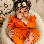 Shadday is registered to the contest to win money with this photo: baby, child, infant, curly_hair, headband, yellow_bow, orange_outfit, sitting, fingers_in_mouth, cozy_chair, soft_furniture, decor, flowers, greenery, sign, milestone, celebration, portrait, indoor, cute