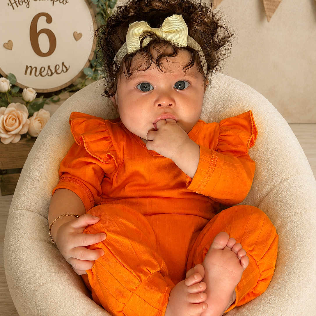 Shadday is registered to the contest to win money with this photo: baby, child, infant, curly_hair, headband, yellow_bow, orange_outfit, sitting, fingers_in_mouth, cozy_chair, soft_furniture, decor, flowers, greenery, sign, milestone, celebration, portrait, indoor, cute