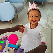 Chelsea participe au concours pour gagner de l'argent avec cette photo : toddler, child, baby, crown, white_dress, smiling, indoor, toy, hands, person, living_room, chair, television, floor, window, curtains, playful, happy, support, footwear