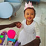 toddler, child, baby, crown, white_dress, smiling, indoor, toy, hands, person, living_room, chair, television, floor, window, curtains, playful, happy, support, footwear