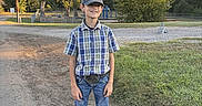 David is registered to the contest to win money with this photo: boy, child, outdoor, plaid_shirt, jeans, boots, baseball_cap, grass, dirt_road, trees, sunlight, smiling, casual_clothing, nature, daytime, young, portrait, standing, rural, scenery
