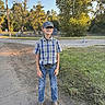 boy, child, outdoor, plaid_shirt, jeans, boots, baseball_cap, grass, dirt_road, trees, sunlight, smiling, casual_clothing, nature, daytime, young, portrait, standing, rural, scenery