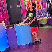 David is registered to the contest to win money with this photo: child, boy, indoor, colorful, mural, playful, curious, tshirt, shorts, footwear, purple_floor, blue_display, red_shorts, black_tshirt, furniture, wall_art, illustration, standing, leaning, looking_up