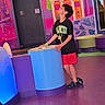 child, boy, indoor, colorful, mural, playful, curious, tshirt, shorts, footwear, purple_floor, blue_display, red_shorts, black_tshirt, furniture, wall_art, illustration, standing, leaning, looking_up