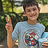 child, boy, smile, peace_sign, mario_kart, tshirt, toy, camera, outdoor, nature, daylight, portrait, happy, casual_clothing, short_hair, greenery, background_blur, person, youth, summer