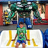 child, boy, smiling, sitting, bench, robot_statue, green_tshirt, minecraft, blue_shorts, outdoor, storefront, sign, advertisement, toy, happy, portrait, daylight, casual_clothing, urban, public_place