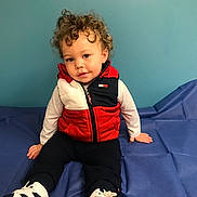 Cameron is registered to the contest to win money with this photo: toddler, child, curly_hair, blue_wall, red_vest, white_long_sleeve, black_pants, sneakers, indoor, sitting, floor_covering, casual_clothing, portrait, person, cute, expression, looking_at_camera, small_earring, young_child