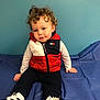 toddler, child, curly_hair, blue_wall, red_vest, white_long_sleeve, black_pants, sneakers, indoor, sitting, floor_covering, casual_clothing, portrait, person, cute, expression, looking_at_camera, small_earring, young_child