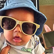 Elijah is registered to the contest to win money with this photo: baby, sunglasses, hat, blue_hat, dinosaur_onesie, reflection, person_reflection, hand, indoor, monitor, headphones, pink_headphones, cute, chubby_cheeks, child, portrait, closeup, clothing, accessory, infant