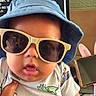baby, sunglasses, hat, blue_hat, dinosaur_onesie, reflection, person_reflection, hand, indoor, monitor, headphones, pink_headphones, cute, chubby_cheeks, child, portrait, closeup, clothing, accessory, infant