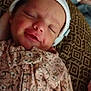baby, blanket, child, cozy, cute, floral_clothing, human, indoor, infant, newborn, onesie, patterned_cushion, peaceful, portrait, resting, sleeping, smile, soft_lighting, white_cap, young_child