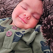Maé participe au concours pour gagner de l'argent avec cette photo : baby, sleeping, smile, face, infant, cozy, blanket, fur, green_clothing, buttoned_shirt, cute, peaceful, portrait, child, soft_texture, resting, indoors, warm, closeup, adorable