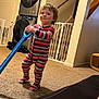 appliance, baby_gate, blue_toy, carpet, child, child_playing, face, floor, furniture, handrail, home_interior, indoor, laundry_machines, person, smiling, staircase, striped_pajamas, toddler, toy, yellow_toy_container