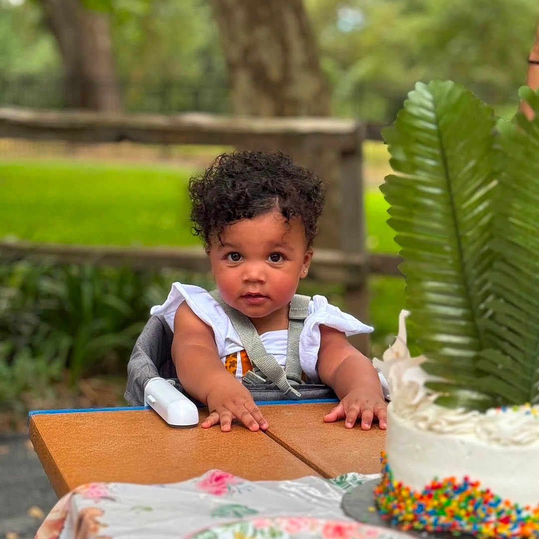 Zahmiyah Frison is registered to the contest to win money with this photo: child, toddler, high_chair, outdoor, table, cake, plate, greenery, tree, curly_hair, food, party, celebration, nature, daylight, baby, face, person, summer, curious