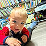Jake is registered to the contest to win money with this photo: baby, infant, child, smile, blue_eyes, bookshelf, books, toys, toy_car, stroller, playroom, floor, wood_floor, red_sleeves, hand, face, portrait, clothing, person, crawling