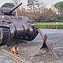 tank, dog, parking_lot, trees, memorial, historic_monument, outdoor, grass, hedge, metal, weapon, canine, quiet, nature, sign, pavement, cloudy_sky, military, vehicle, museum