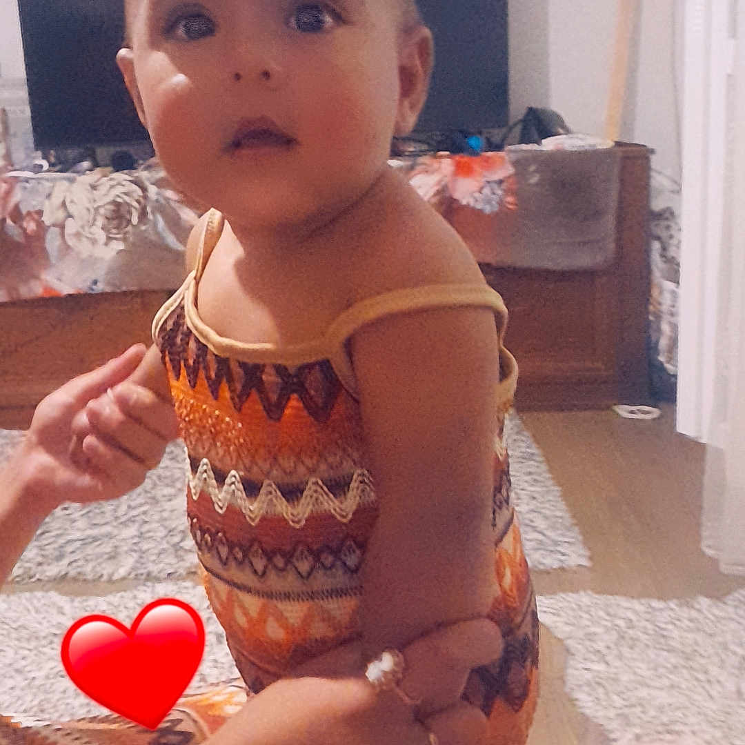 Swahili a rejoint le concours — aidez-le/la à gagner de superbes lots ! adult_hand, carpet, child, curious, dress, eyes, face, furniture, head, holding, home, indoor, living_room, orange, pattern, person, room, skin, television, toddler