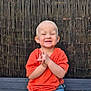toddler, child, smiling, orange_shirt, blue_jeans, bench, outdoor, happy, hands_clasped, portrait, casual_clothing, short_hair, fence, wooden_bench, daylight, cute, young_child, sitting, person, joyful
