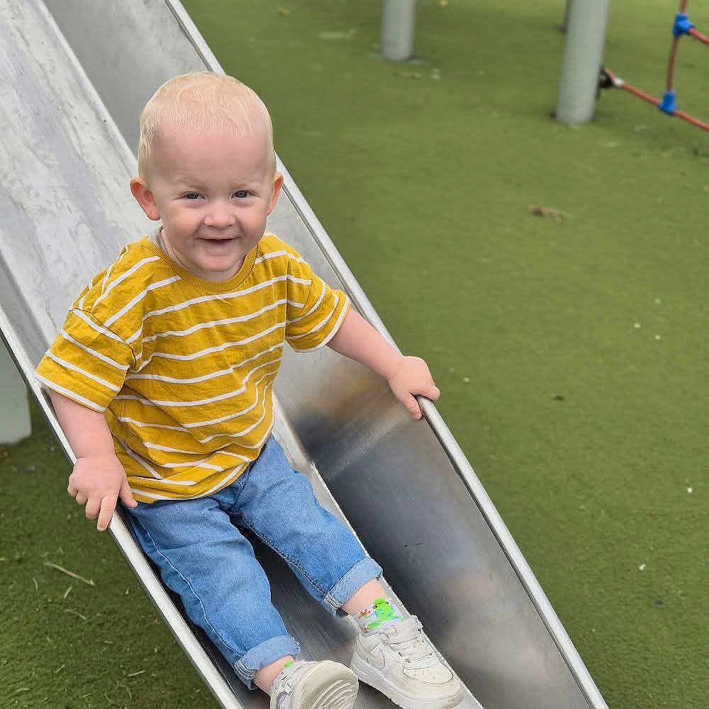 Carson is registered to the contest to win money with this photo: child, toddler, slide, playground, yellow_shirt, striped_shirt, jeans, sneakers, smiling, outdoor, metal_slide, green_ground, happy, fun, playing, casual_clothing, young_child, daylight, recreation, park