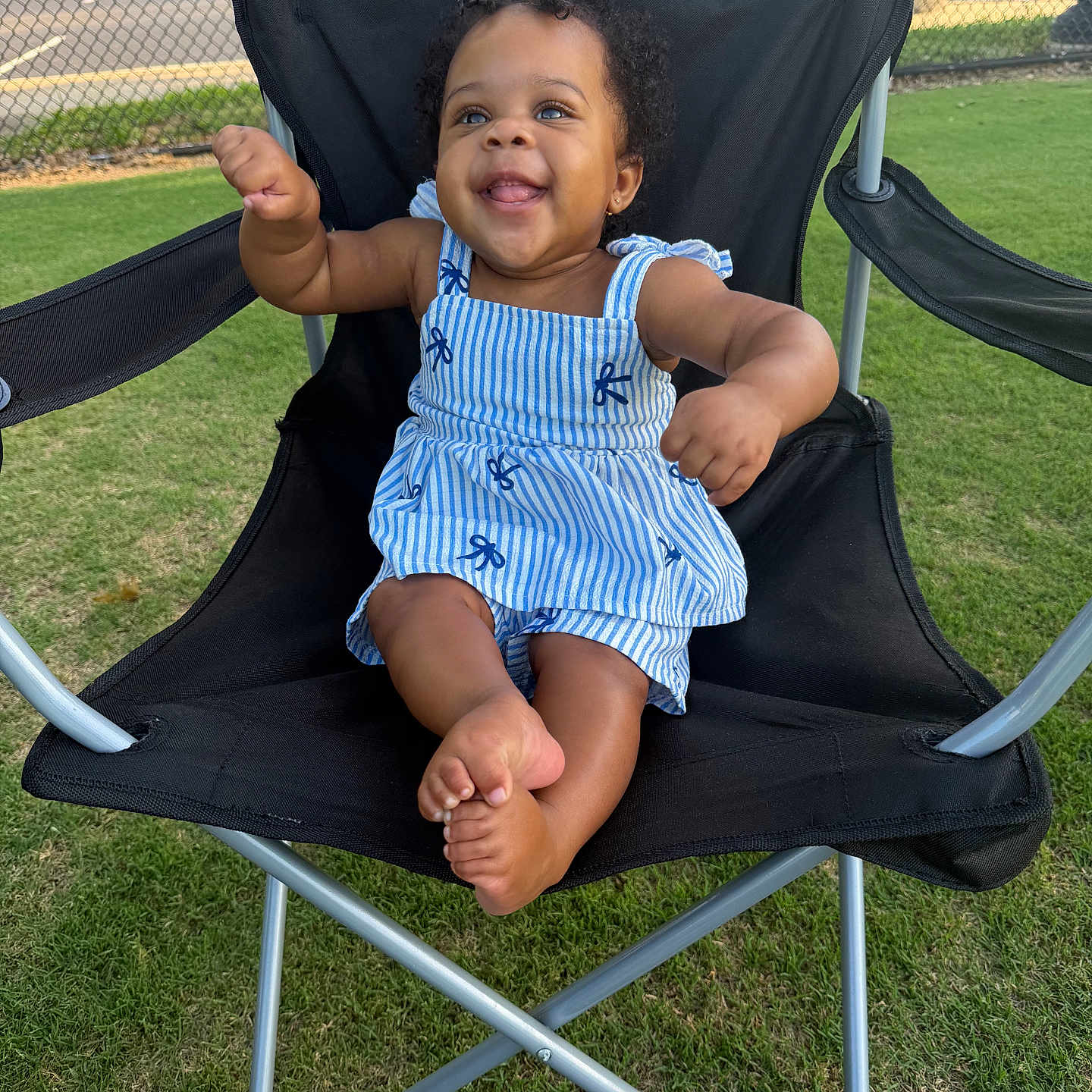Davina is registered to the contest to win money with this photo: baby, child, smiling, blue_dress, striped, chair, outdoor, grass, happy, cute, sitting, black_chair, summer, sunny, portrait, infant, playful, person, curly_hair, barefoot