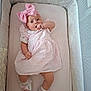 baby, crib, pink_bow, dress, shoes, socks, infant, child, cute, lying_down, quilted_padding, thumb_sucking, headband, striped_clothing, indoor, soft_lighting, white, comfortable, looking_at_camera, adorable