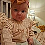 baby, child, headband, bow, bed, bedroom, lamp, blanket, pattern, sitting, clothing, long_sleeve, pants, indoor, person, cute, face, blue_eyes, hand, soft_lighting