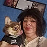 woman, person, dog, chihuahua, pet, selfie, portrait, indoors, blue_wall, framed_art, close_up, face, tongue_out, brown_dog, black_shirt, necklace, smiling, companion, casual, photography