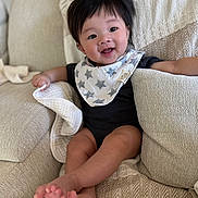 Ray is registered to the contest to win money with this photo: baby, infant, child, smiling, bib, star_bib, onesie, living_room, couch, blanket, pillow, hair, eyes, cheeks, toes, legs, feet, portrait, happy, soft_texture