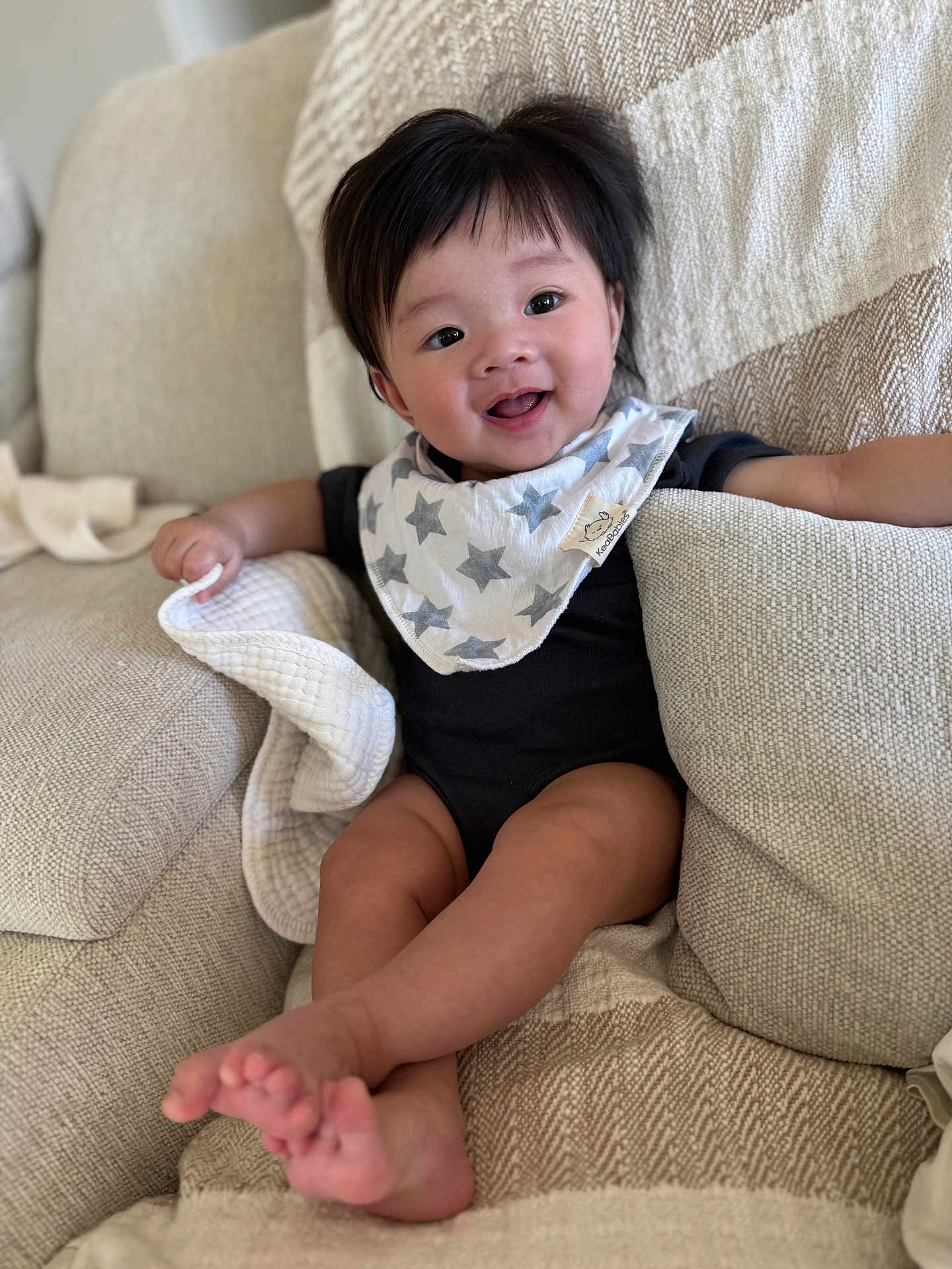 Ray is registered to the contest to win money with this photo: baby, infant, child, smiling, bib, star_bib, onesie, living_room, couch, blanket, pillow, hair, eyes, cheeks, toes, legs, feet, portrait, happy, soft_texture