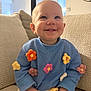 baby, child, smiling, blue_sweater, knitted_flowers, sitting, couch, indoor, happy, portrait, cozy, cute, infant, warm_light, furniture, person, casual_clothing, home, small_child, cheerful