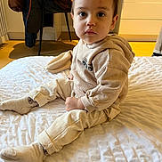 Elio participe au concours pour gagner de l'argent avec cette photo : toddler, child, indoor, bed, white_bedspread, beige_clothing, socks, face, expression, curious, chair, jacket, wooden_floor, modern_kitchen, person, baby, sitting, hoodie, pants, cozy