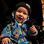 baby, child, smiling, blue_onesie, beanie, stroller, indoor, happy, clothing, infant, cute, face, person, seatbelt, warm_clothing, fleece, portrait, hands, young_child, cozy