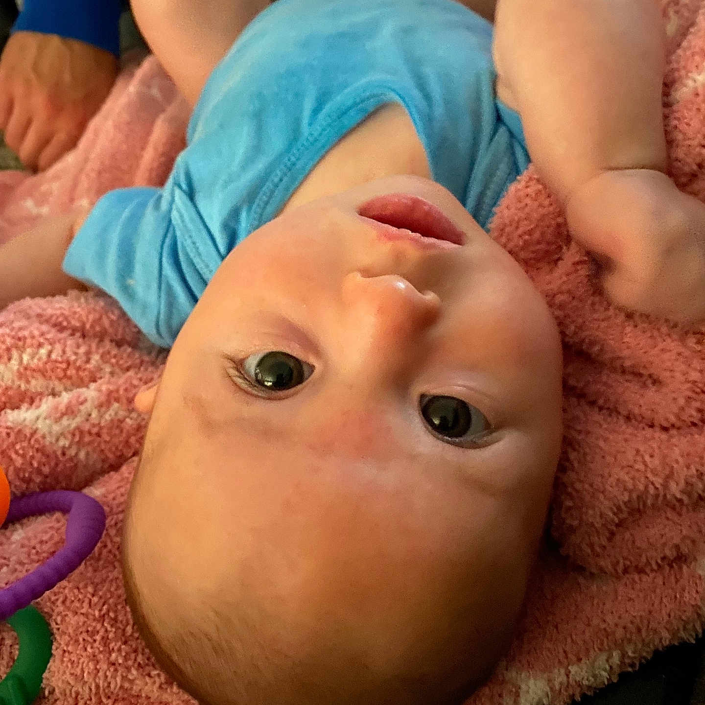 Skadi is registered to the contest to win money with this photo: baby, blue_onesie, child, closeup, curious, eyes, face, fingers, hand, head, indoor, infant, lying_down, mouth, person, pink_towel, relaxed, skin, soft_texture, toys