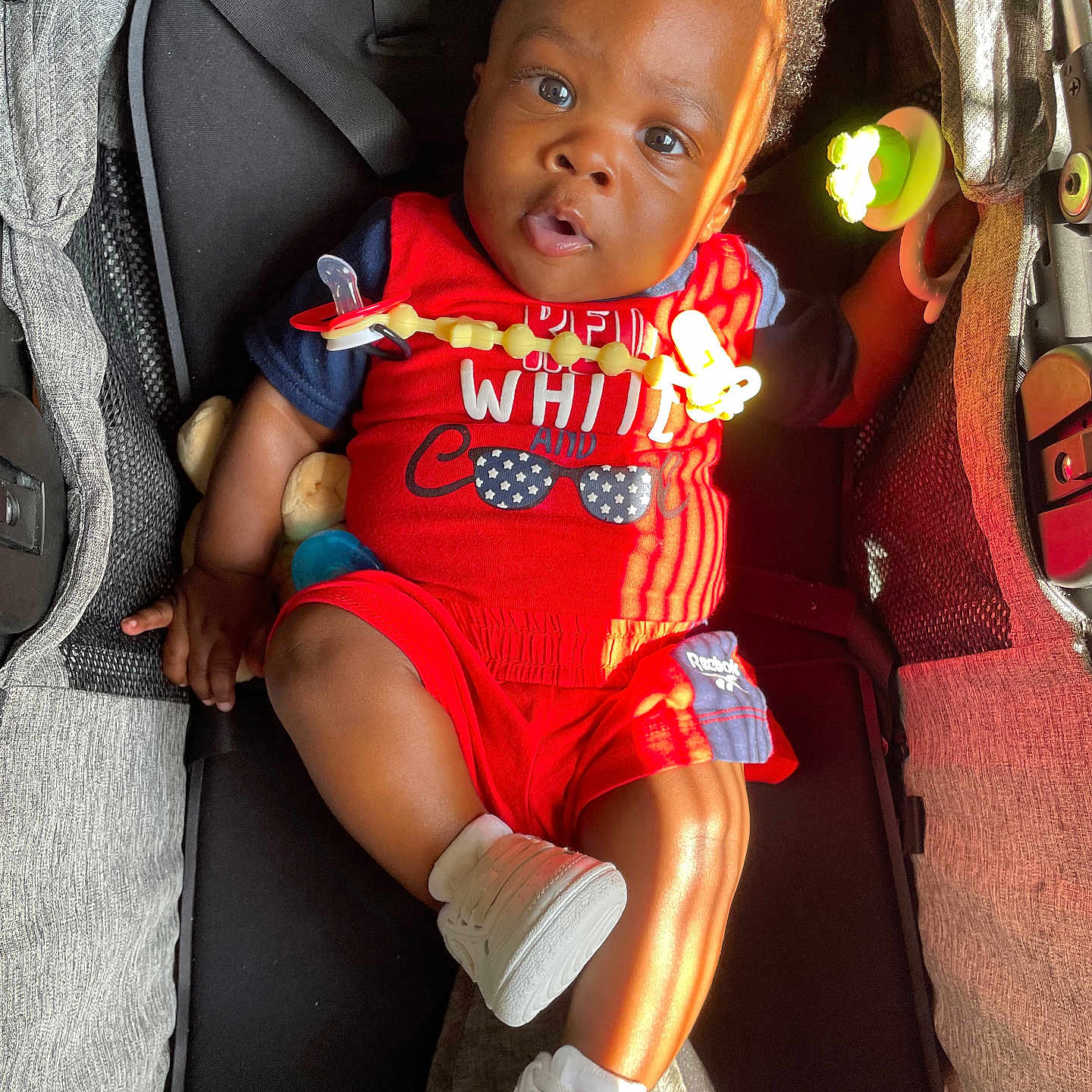 Jayveon is registered to the contest to win money with this photo: baby, black_hair, bright, casual, child, comfortable, curly_hair, cute, indoor, infant, pacifier_clip, portrait, red_clothing, relaxed, seatbelt, smiling, stroller, sunlight, toy, white_shoes