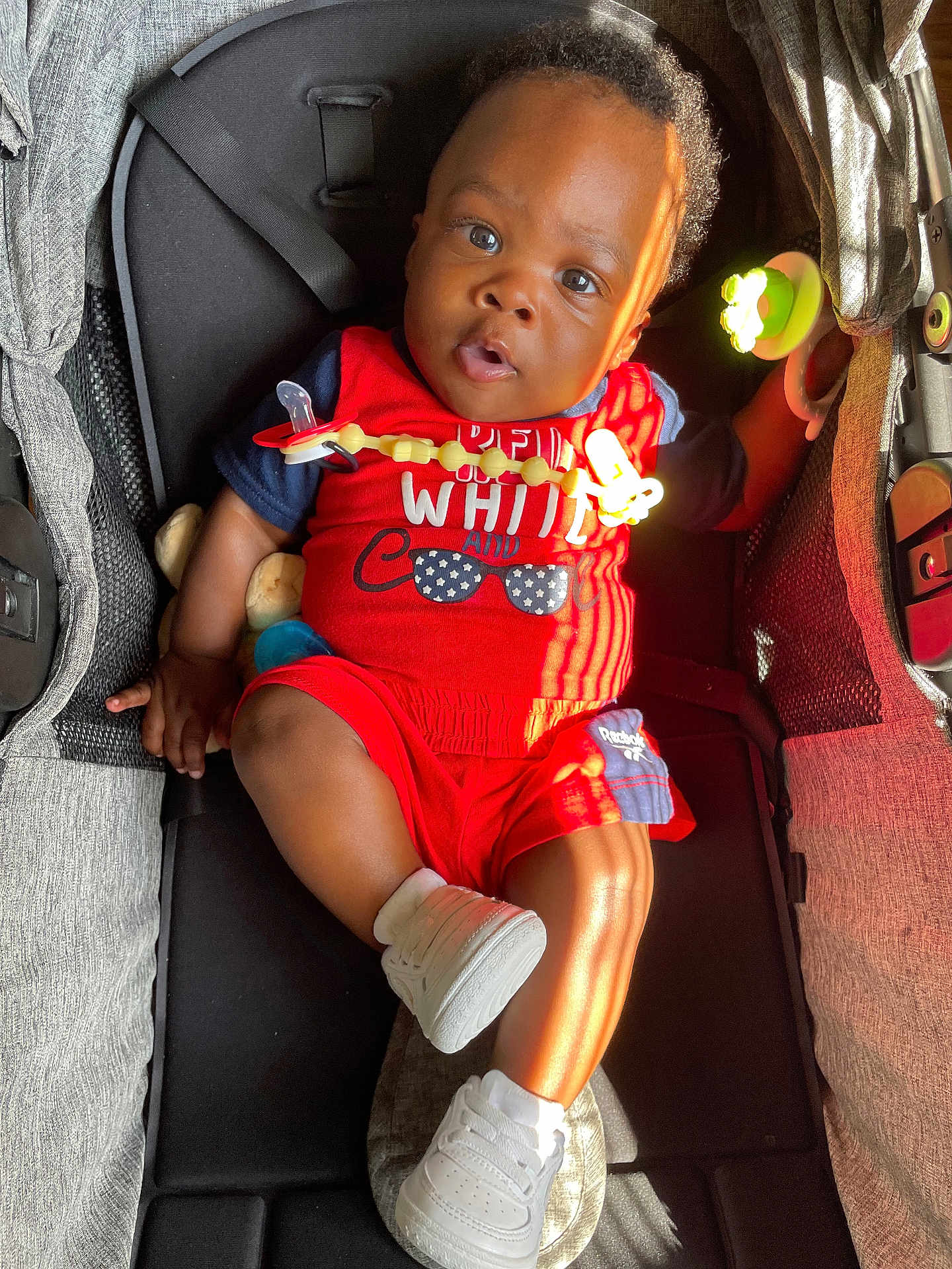 Jayveon is registered to the contest to win money with this photo: baby, child, stroller, red_clothing, white_shoes, pacifier_clip, sunlight, indoor, cute, infant, portrait, casual, smiling, relaxed, toy, seatbelt, curly_hair, black_hair, bright, comfortable