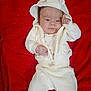 baby, infant, white_clothing, hoodie, blanket, red_background, cute, lying_down, small_hands, face, portrait, soft_fabric, warm_clothes, child, newborn, expression, indoors, cozy, casual, sleepwear