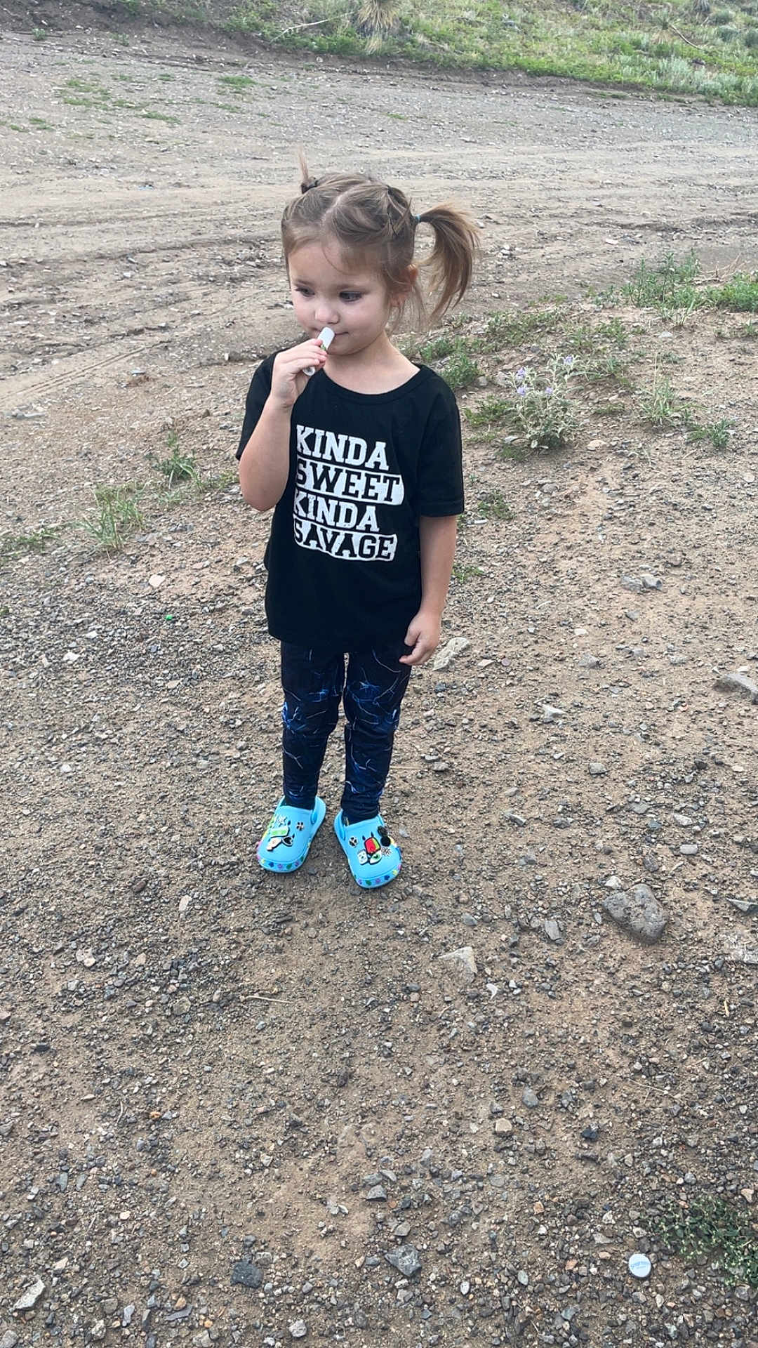 Mila is registered to the contest to win money with this photo: child, girl, pigtails, black_shirt, text_on_shirt, blue_shoes, outdoor, dirt_path, rocks, grass, standing, casual_clothing, young_child, nature, expression, person, footwear, daylight, playful, cute