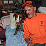 man, dog, bed, blue_sheet, orange_shirt, orange_hat, indoor, clutter, furniture, pillow, pet, canine, casual, smiling, flash_reflection, beard, seated, human, domestic, relaxed