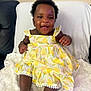 Kayla is registered to the contest to win money with this photo: baby, child, smiling, dress, yellow_dress, pineapple_pattern, sitting, blanket, couch, indoor, happy, cute, curly_hair, foot, hand, portrait, infant, person, furniture, white_blanket