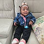 baby, child, denim_jacket, pacifier, bow, pink_shoes, polka_dots, black_pants, leather_couch, cushion, floral_pillow, smiling, seated, indoors, cute, happy, person, portrait, small_hands, curly_hair
