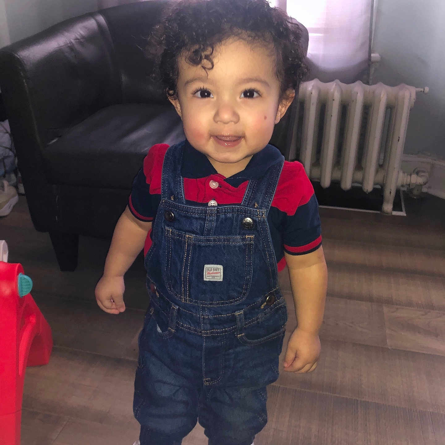 Romeo joined the competition — help win amazing prizes! toddler, child, smiling, curly_hair, denim_overalls, red_shirt, white_shoes, indoor, armchair, radiator, window, wood_floor, toy, happy, person, standing, face, clothing, home, casual