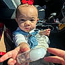 child, toddler, red_bow, denim_overalls, white_shirt, car_interior, car_seat, cup_holder, hand, face, curious, infant, seatbelt, sunlight, window_light, baby, person, close_up, expression, reaching