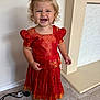 barefoot, blonde_hair, carpet, child, curly_hair, cute, face, happy, home, indoor, person, playful, portrait, power_cord, red_dress, smiling, standing, toddler, traditional_clothing, wall