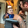 Kayce joined the competition — help win amazing prizes! baby, high_chair, man, feeding, tattoo, smile, indoor, deer_mount, wood_floor, door, lamp, shadow, glasses, shorts, wall, person, child, happy, meal, spoon