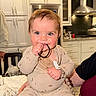 baby, child, kitchen, onesie, hair_tie, chewing, curious_eyes, cute, indoor, modern_kitchen, white_cabinets, stove_hood, person, infant, playing, seated, pacifier_clip, toy, smiling, hand