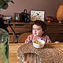 toddler, child, table, bowl, snack, face, chair, wooden_table, placemat, glass, bottle, home_decor, pumpkin, clock, handbag, curious, indoor, brown_wall, plant, seated